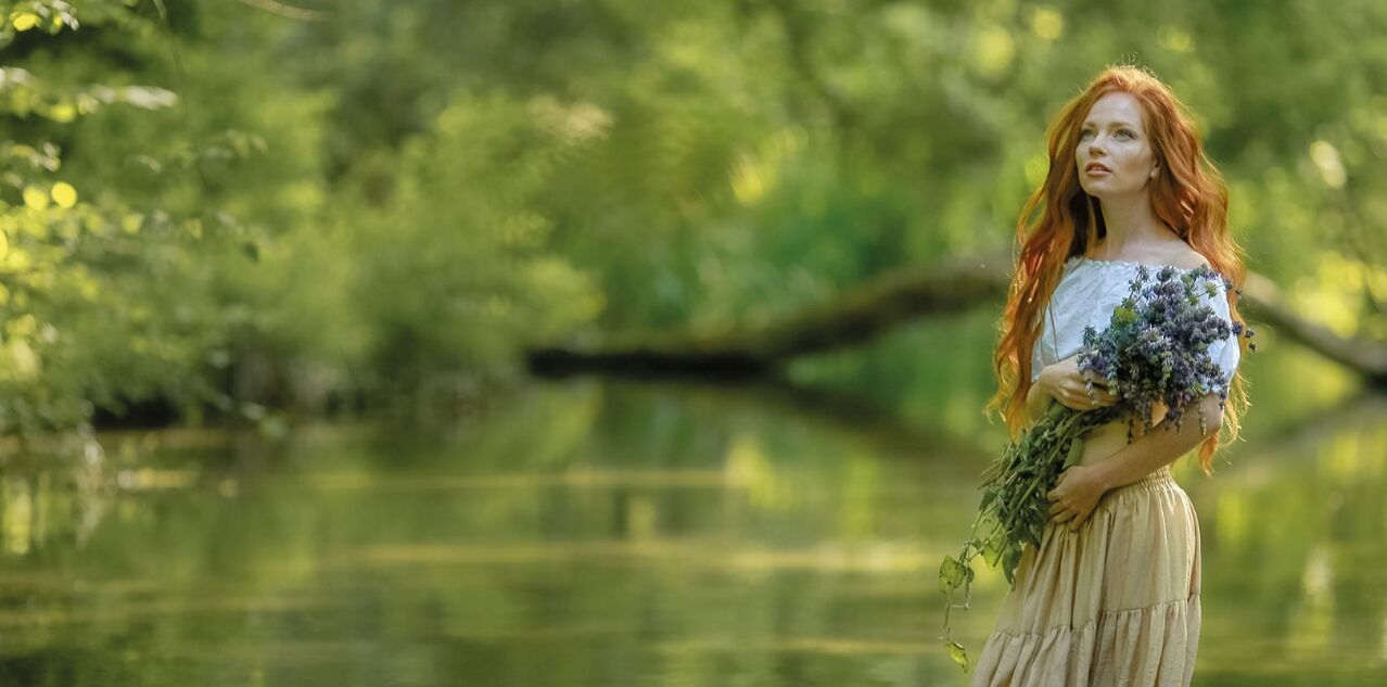 Eine Frau mit roten, langen Haaren in einem Kleid steht in einem grünen Wald mit einem trockenen Blumenstrauß in der Hand.