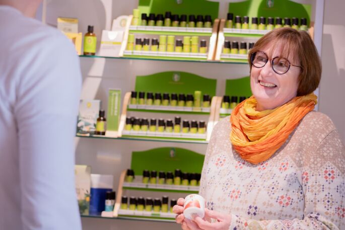 Die Inhaberin Frau Doris Mittelstädt lächelt zu einem Kunden und hält eine Creme in der Hand. Hinter ihr ein Regal mit Primavera Ölen.