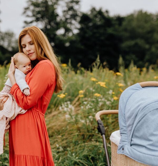 Eine Frau in einem roten, langen Kleid trägt ein kleines Baby auf dem Arm auf einem Feldweg. Rechts steht ein hellblauer Kinderwagen.