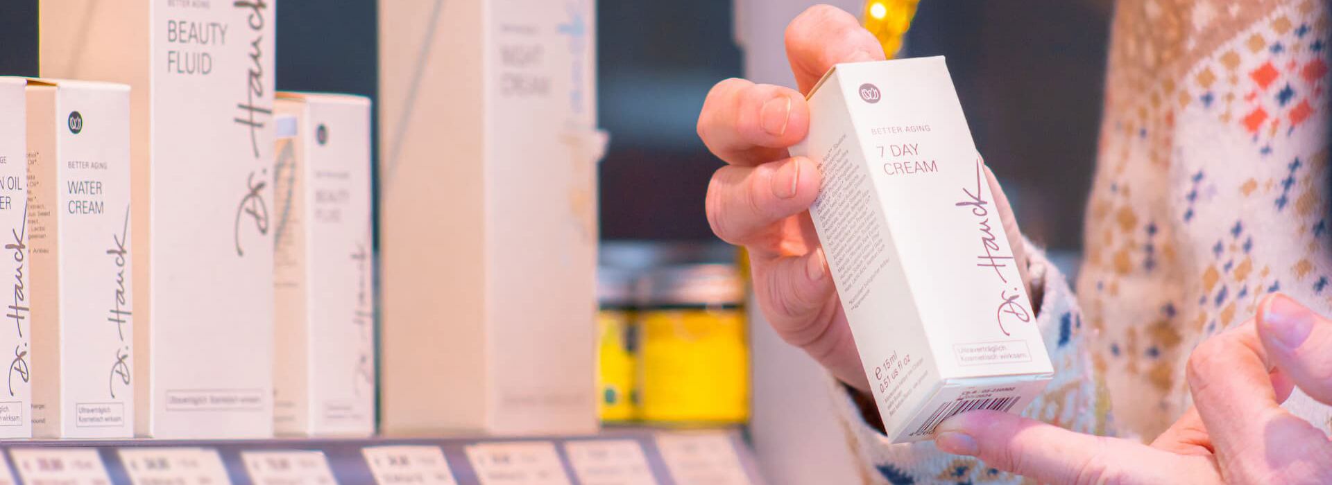 Entdecken Sie Dr. Hauschka Naturkosmetik bei Hautnah Naturwaren. Zwei Hände halten ein Dr. Hauschka Produkt. Im Hintergrund ein Regal mit Dr. Hauschka Produkten.