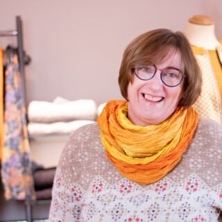 Entdecken Sie Naturbekleidung bei Hautnah Naturwaren. Die Inhaberin Frau Mittelstädt trägt einen gelben Loopschal und lächelt in die Kamera. Hinter ihr befinden sich Regale und Kleiderstangen mit Kleidung.