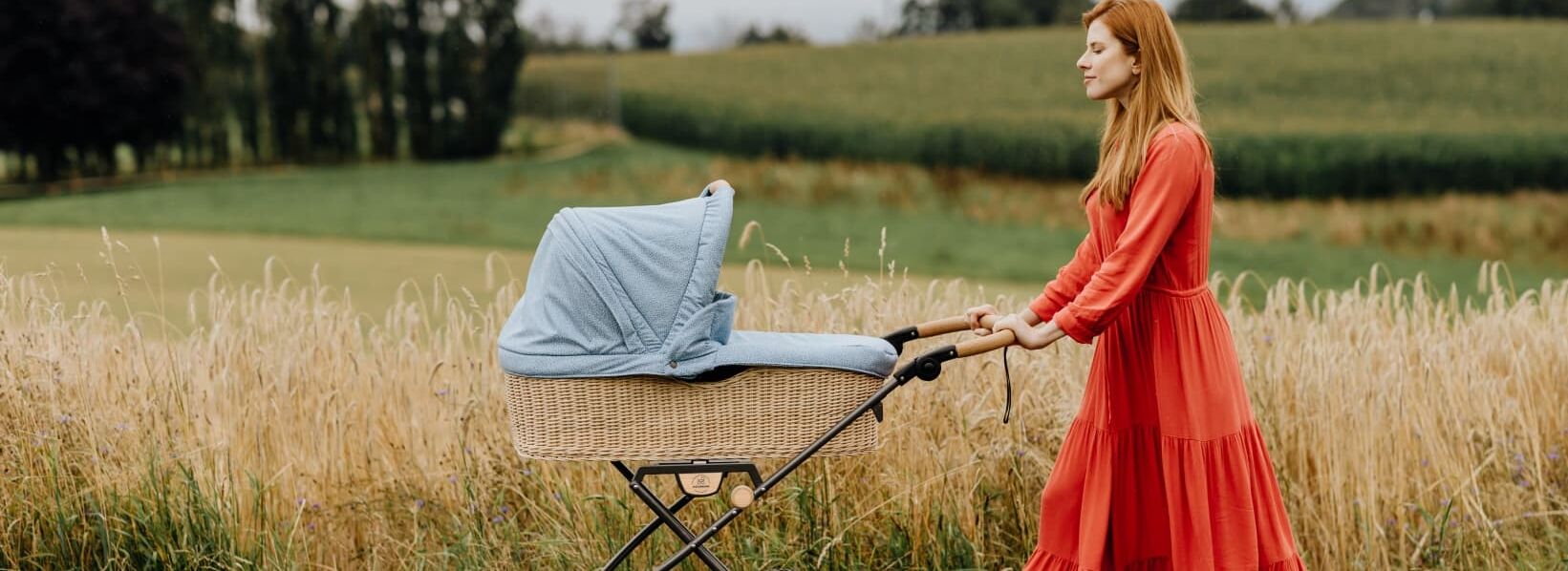 NATURKIND Kinderwagen Ida - erhältlich bei Hautnah Naturwaren Eine Frau in einem roten, langen Kleid fährt ihr Baby in einem Kinderwagen auf einem Feldweg spazieren.