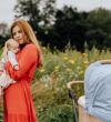 NATURKIND Kinderwagen Ida - erhältlich bei Hautnah Naturwaren Eine Frau in einem roten, langen Kleid trägt ein kleines Baby auf dem Arm auf einem Feldweg. Rechts steht ein hellblauer Kinderwagen.